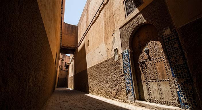 A narrow and evocative alley in the medina of Meknes, with ochre-colored walls and an ancient wooden door decorated with blue mosaics.