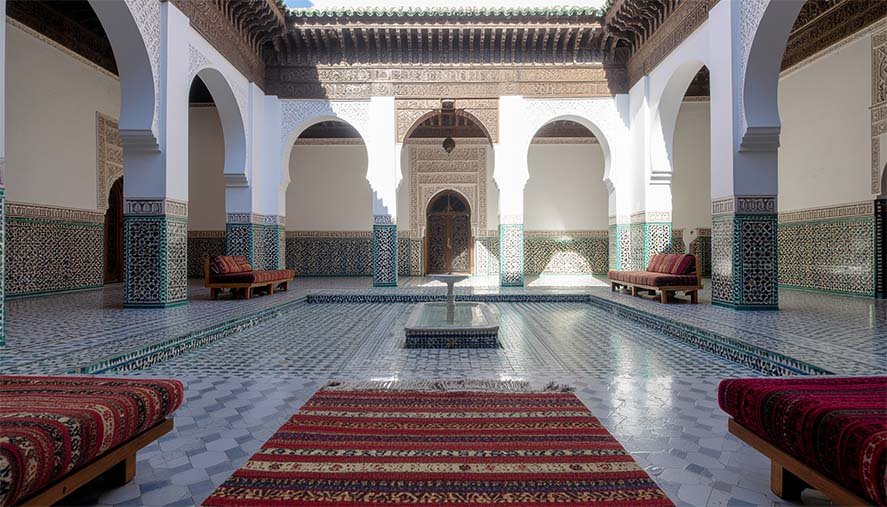 Interior of a Moroccan riad with traditional architecture, ceramic decorations, and colorful cushions arranged on sofas, with a fountain in the center.