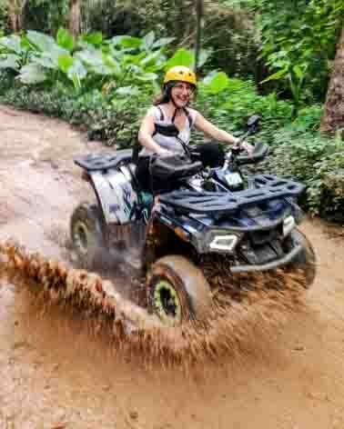 A smiling woman in a yellow helmet has fun riding an ATV and splashing through a large mud puddle on a jungle trail.