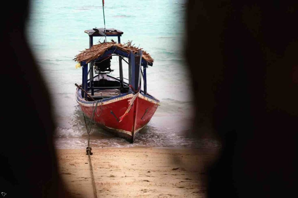 A traditional red and blue Thai long-tail boat with a thatched roof rests on the shore of a sandy beach in Phuket.