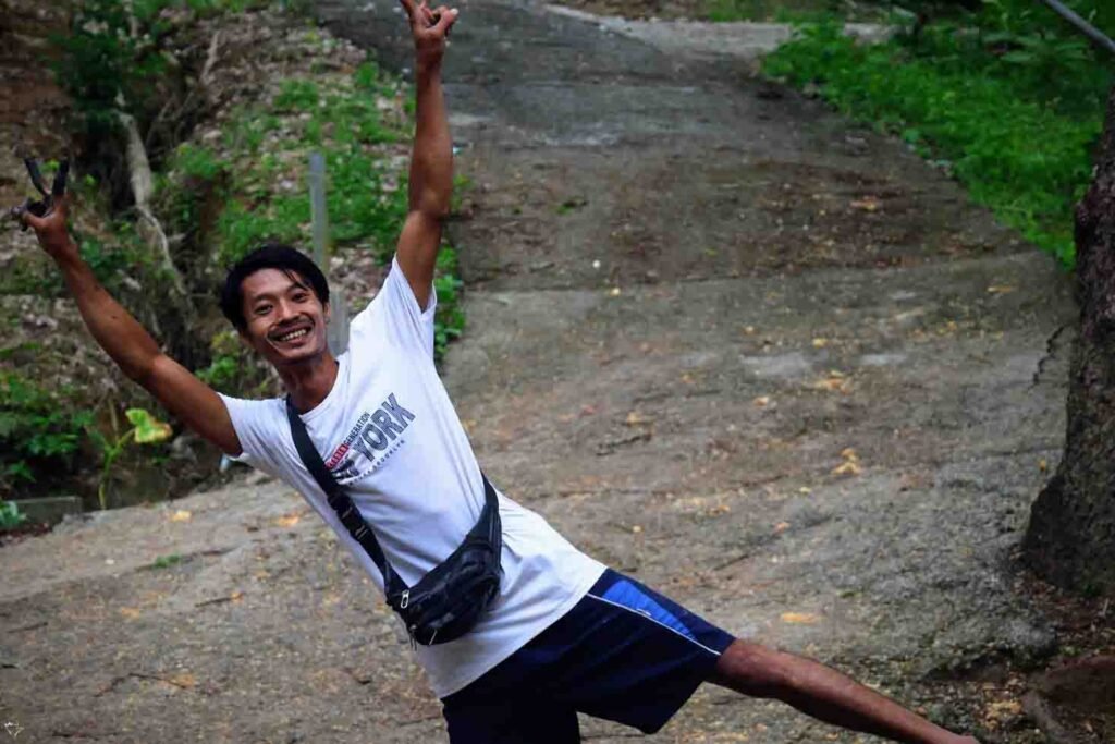 A happy Thai man in a white shirt smiles and makes peace signs with both hands while posing on a path in a lush, green area.