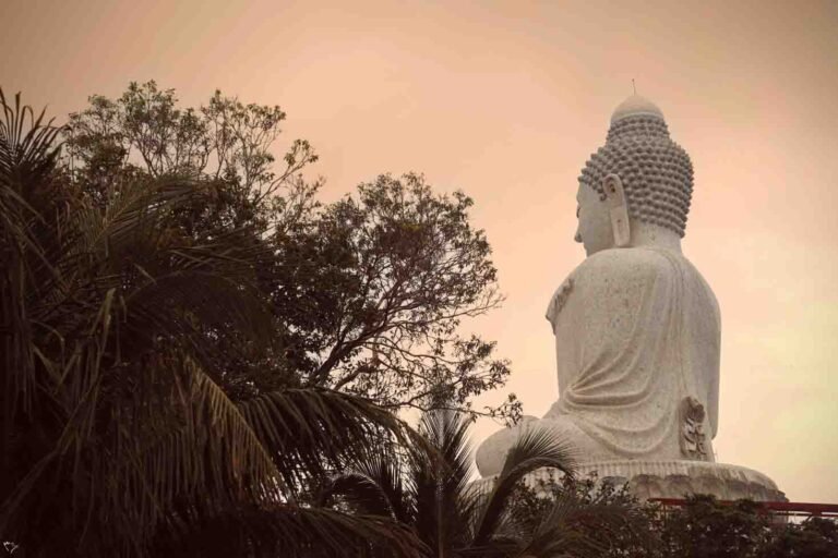 The back of the large white marble Big Buddha statue in Phuket, seen from below and surrounded by trees against a warm-toned sky.