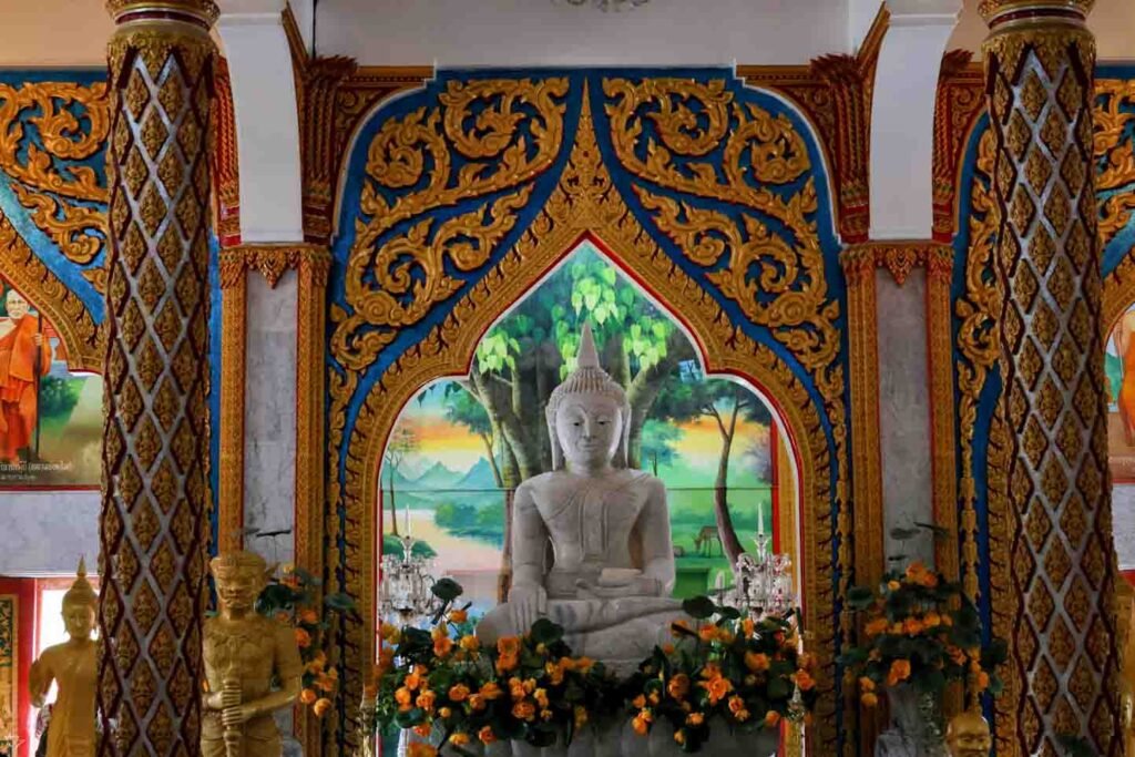 An ornate Thai temple interior with a white Buddha statue at the center, surrounded by flowers and intricate gold and blue decorations.